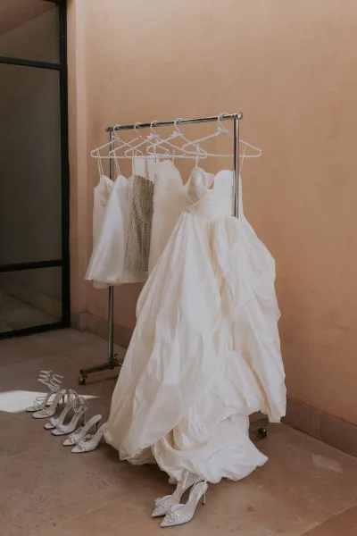 Wedding dress on a clothing rack with pearl hangers, shown with rhinestone heels beside a stucco wall window in a hallway