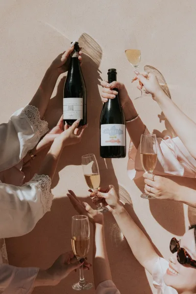 Wedding champagne toast as bridesmaids in lace and satin robe sleeves clink flutes, engagement ring sparkling against a sunlit stucco wall