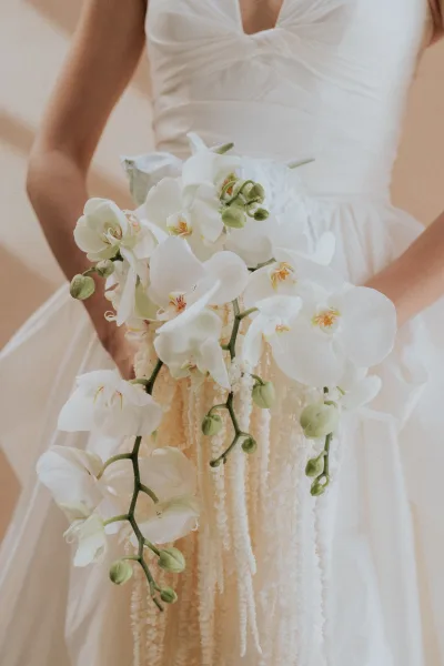 Bridal bouquet of white orchid bouquet with long cascading stems held against a strapless textured wedding dress indoors by a neutral wall