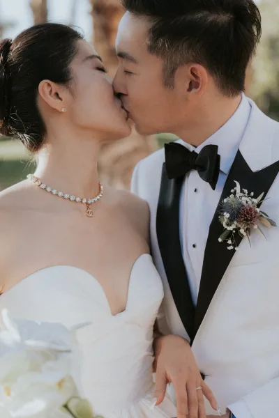 Wedding kiss portrait of bride and groom kissing close up, her diamond necklace and rings catching light amid sunlit greenery bokeh