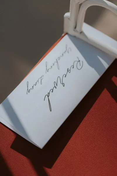 Wedding place card with calligraphy place cards on a white tent card tucked into an orange envelope on a chair, set against red fabric in sunlit shadows