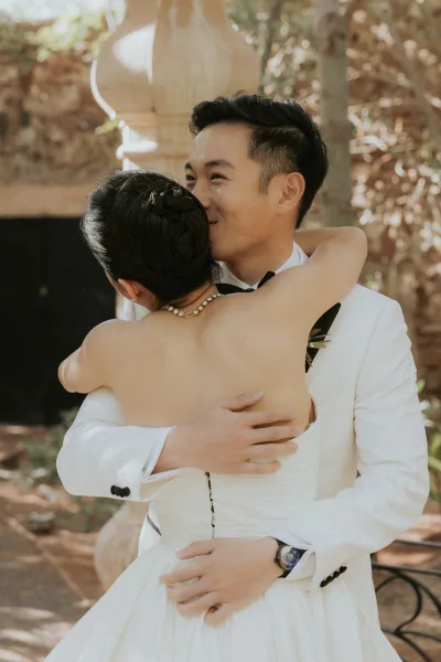 Ceremony hug as bride and groom embrace, his white tuxedo jacket and black bow tie glowing in dappled garden sunlight near stone steps
