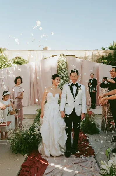 Recessional moment as bride and groom walking down aisle through petal confetti under a greenery arch in an outdoor courtyard with guests cheering