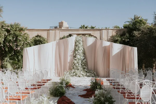 Ceremony setup with draped fabric backdrop and floral installation, white aisle runner with rust fabric in a sunny courtyard under blue sky