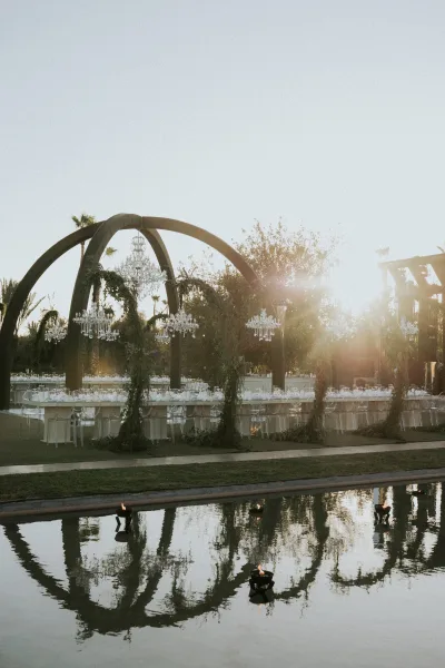 Reception tablescape at an outdoor wedding reception with long banquet tables, chandeliers, greenery garlands, and floating candles by a reflecting pool at sunset