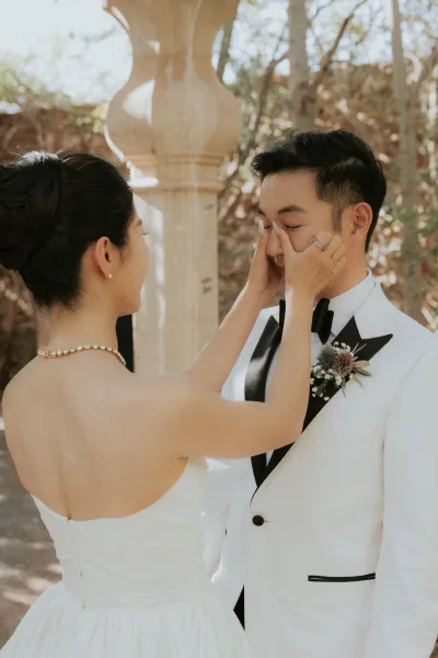 First look moment as bride touches groom’s face while he wipes tears, wearing a white tuxedo and black bow tie in sunlit garden greenery