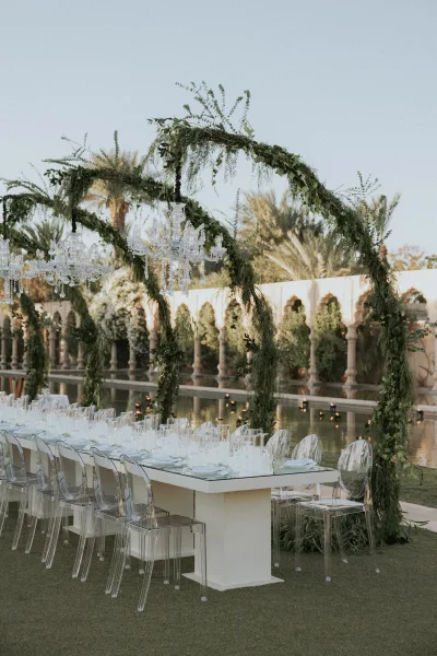 Reception tablescape with an outdoor reception table on a glass dining table beneath crystal chandeliers and hanging greenery by a reflecting pool
