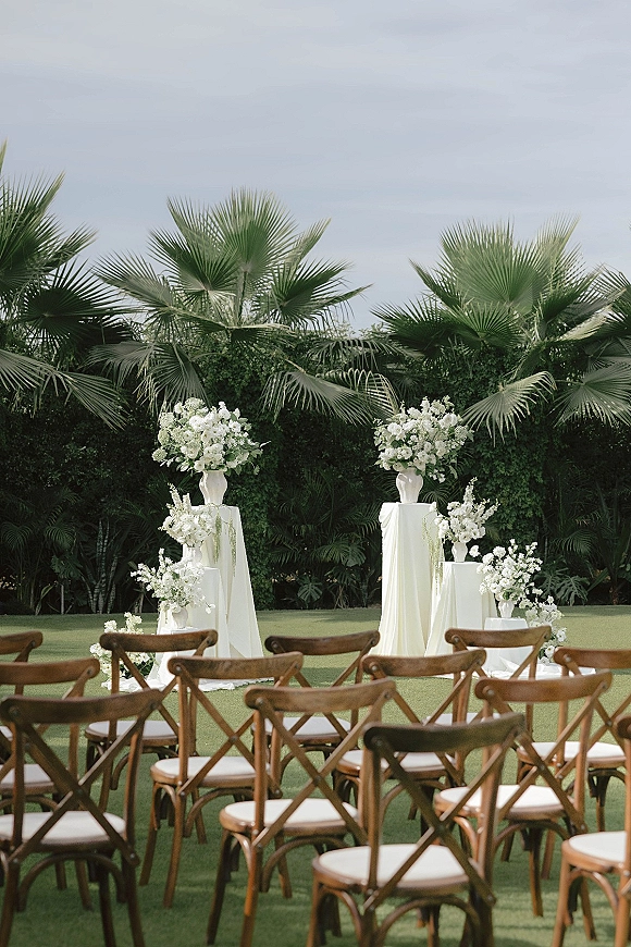 Ceremony setup with outdoor wedding ceremony seating, wooden cross back chairs and white floral pedestal stands on a lawn with palms under cloudy sky