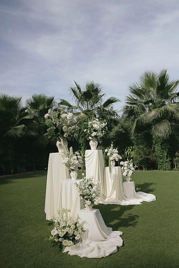 Ceremony altar decor with outdoor wedding altar styling, featuring white floral arrangements on pedestals with draped fabric on a lawn by palms