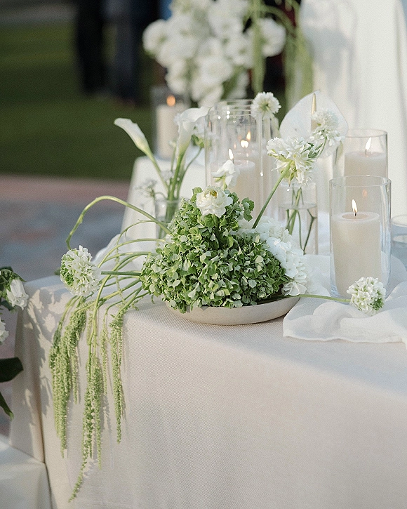 Wedding tablescape with a white wedding centerpiece of calla lilies, greenery, bud vases, and taper candles in glass hurricanes on a lawn