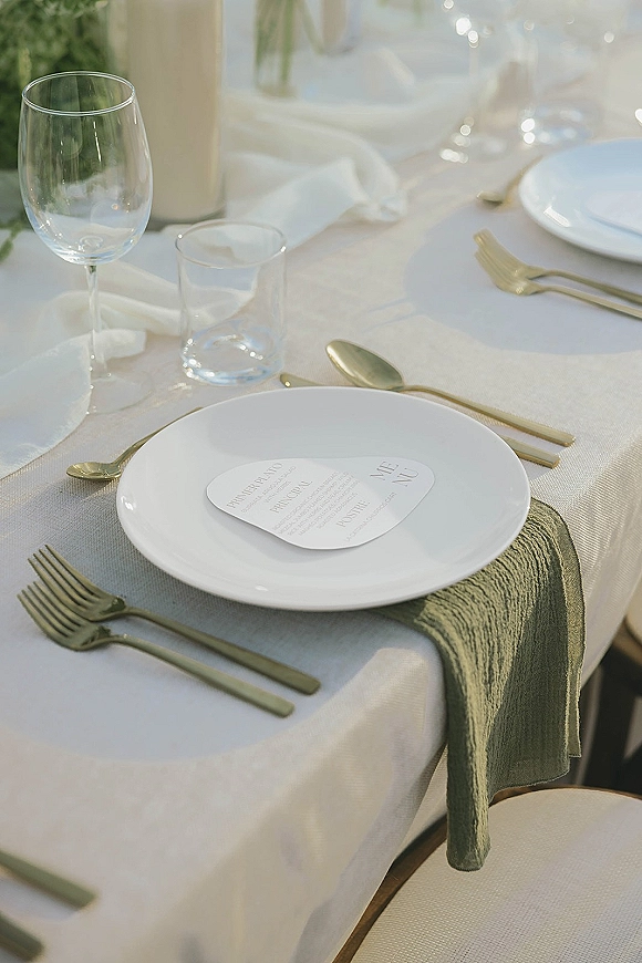 Reception tablescape with wedding place setting featuring white plates, gold flatware, menu card, linen napkin, and sage runner on white cloth