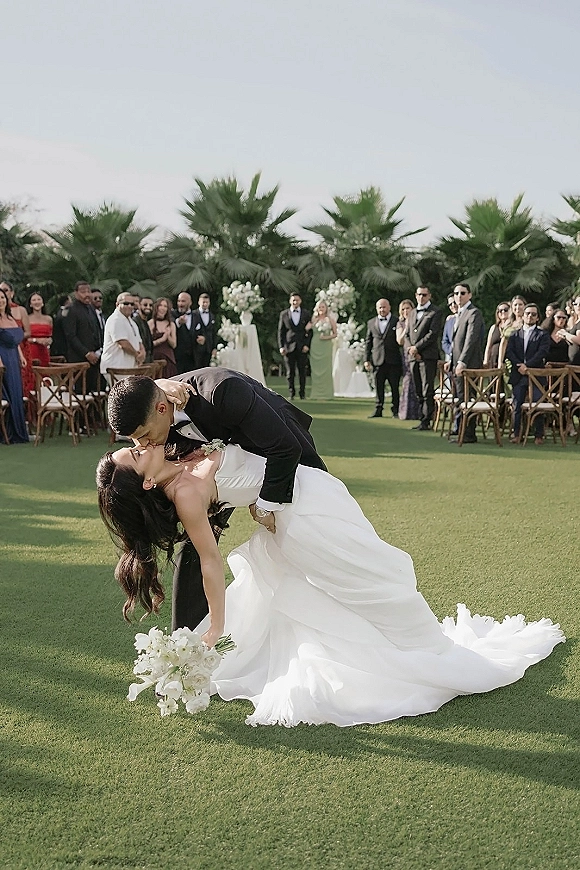 Wedding kiss portrait as bride in strapless gown dips into groom in black tux, bouquet and long train flowing down a palm-lined aisle with guests