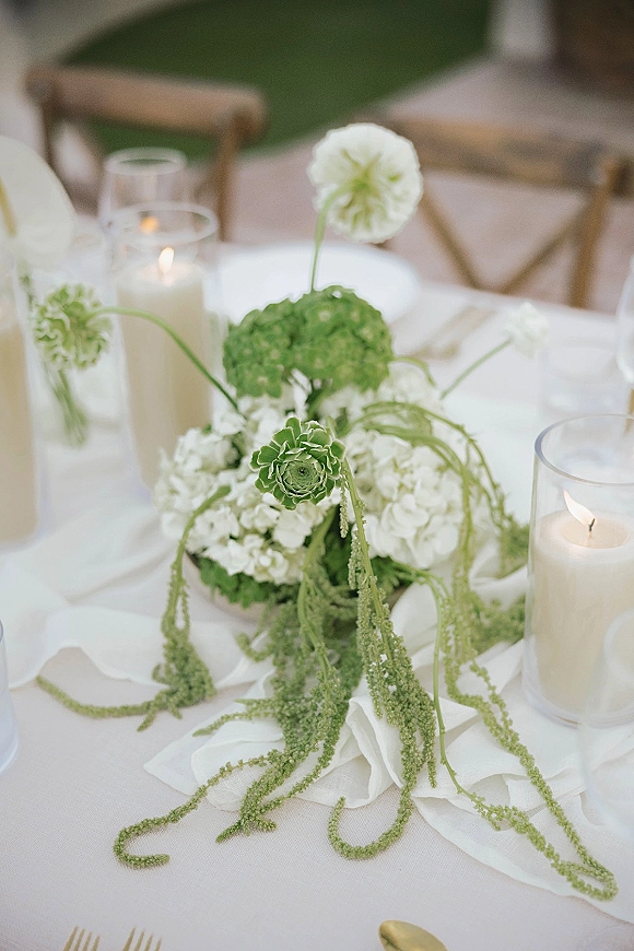 Wedding centerpiece with green and white hydrangeas and trailing amaranthus beside glass cylinder candles on a reception tablescape with gold flatware