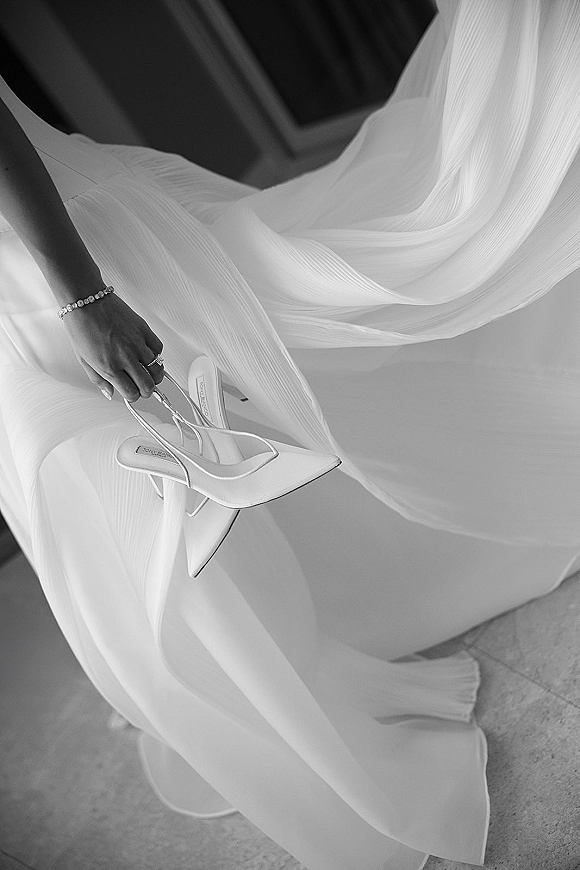 Bridal heels, white strappy heels held in hand beside flowing bridal gown fabric, showing bracelet and engagement ring on a stone floor indoors