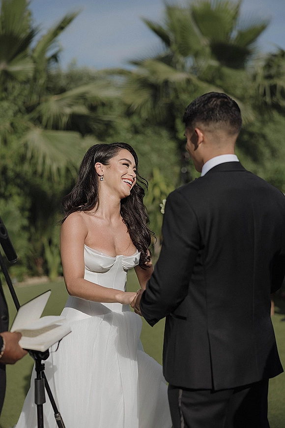 Wedding vows at an outdoor wedding ceremony as bride in a strapless satin gown holds hands with groom in tux under palm trees and blue sky
