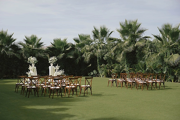 Ceremony setup for an outdoor wedding ceremony with wood cross back chairs and white floral pedestals lining an aisle on a palm-lined lawn