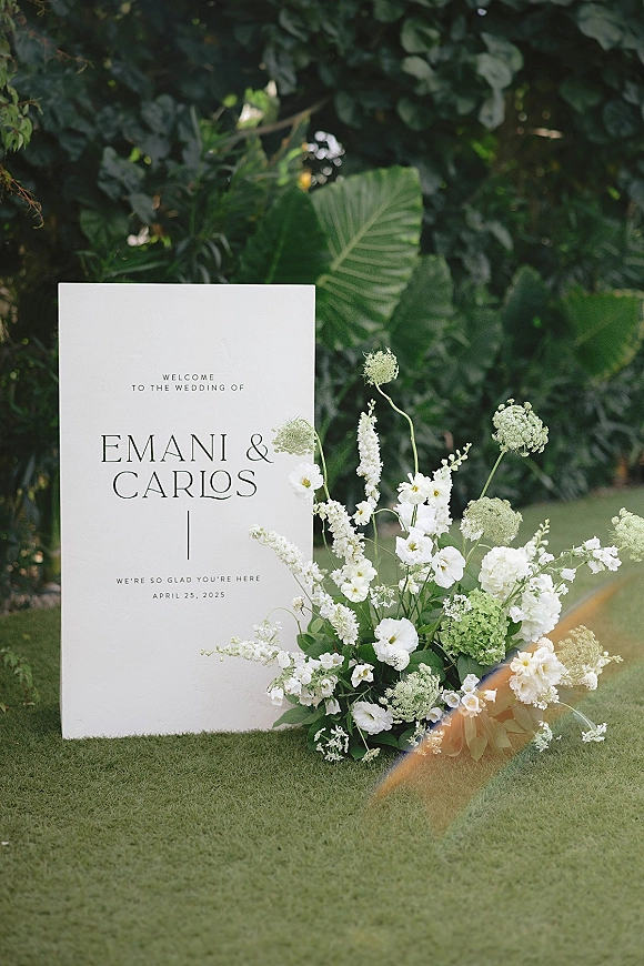 Wedding welcome sign with white and green floral spray at the base, set on a grass lawn in front of a leafy hedge