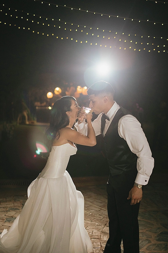Reception moment as newlyweds toast with champagne, bride in strapless gown and groom in vest under string lights on a patio at night