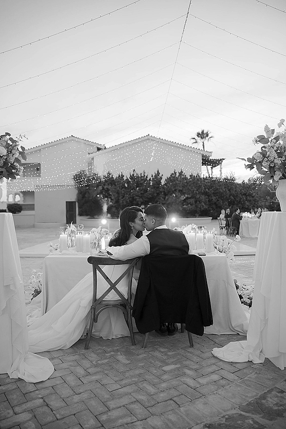 Wedding kiss portrait of newlyweds seated at a candlelit sweetheart table under string lights in a courtyard patio, bride’s train draped behind