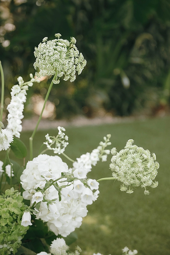 Wedding florals with white wedding flowers and lush greenery arranged on the ground, set against garden foliage, lawn, and trees