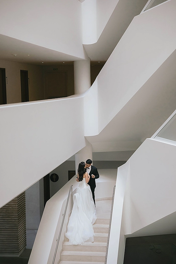 Wedding couple portrait of bride and groom on stairs, her strapless dress with long train flowing in a minimalist modern white stairwell