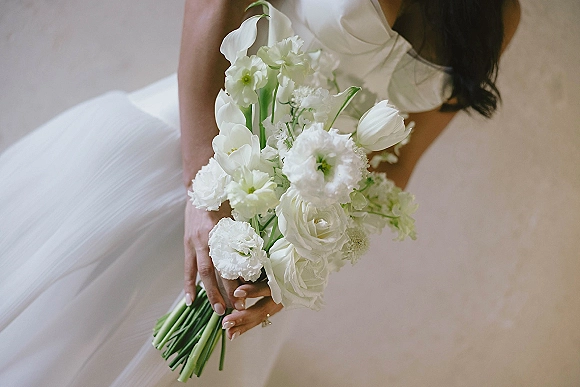 Bridal bouquet, white wedding bouquet of tulips, roses, and lisianthus held against a bridal gown, engagement ring visible on hand