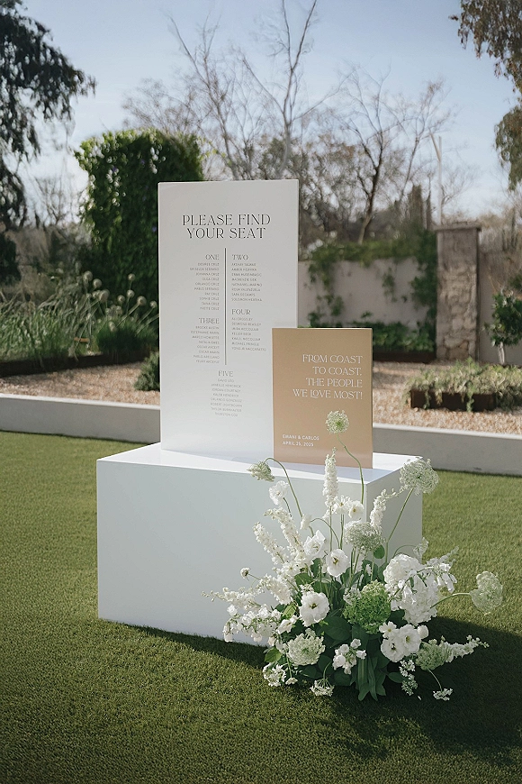 Wedding seating chart display on a white pedestal with white and green floral accents in an outdoor garden with stone wall and blue sky