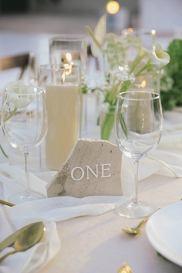 Reception tablescape with wedding table decor, stone table number, cylinder candles, white florals and greenery, gold flatware on white linens