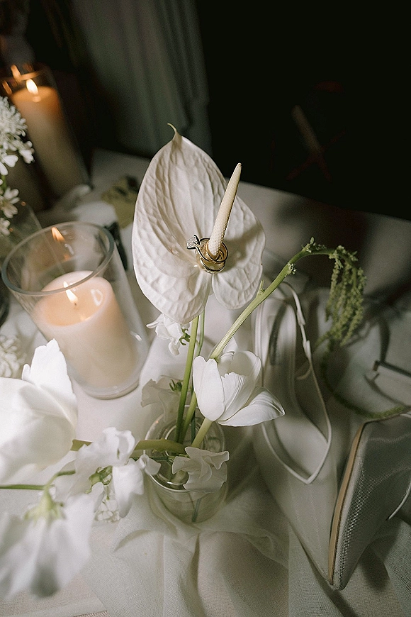 Wedding rings detail with engagement ring and band on white anthurium, beside tulips, ribbon, clutch and glowing hurricane candles on a dim tabletop