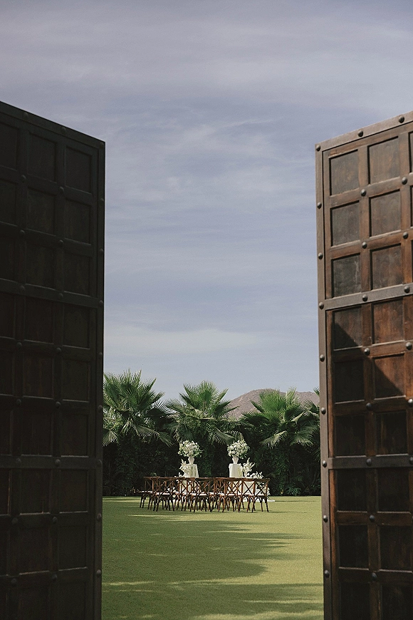 Ceremony setup with wooden chairs and floral pedestals facing a fabric-draped altar by large doors, with palms and mountains beyond
