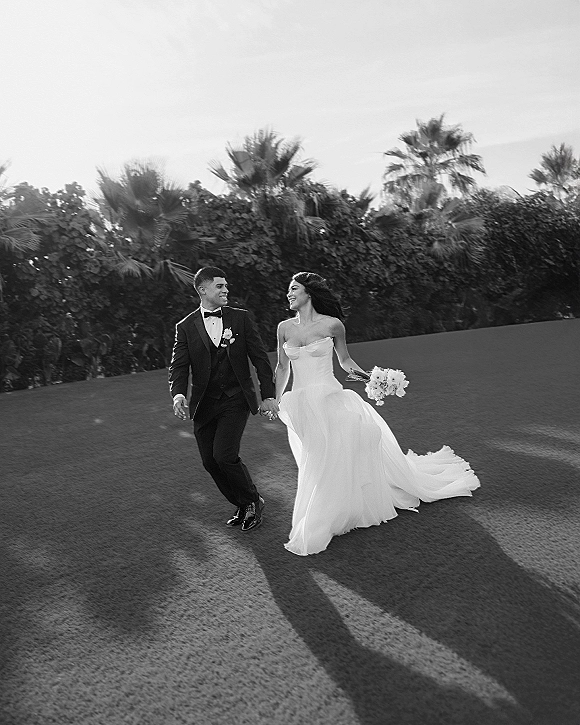 Wedding couple portrait in black and white, bride in strapless dress holding bouquet and groom in tuxedo walking on a palm-lined lawn