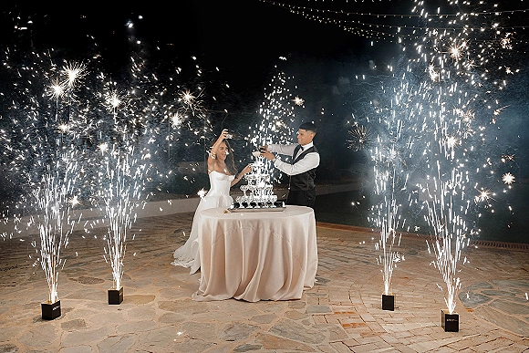 Champagne tower moment as bride in strapless gown and groom in black vest pour bubbles amid sparklers and string lights on a stone patio at night