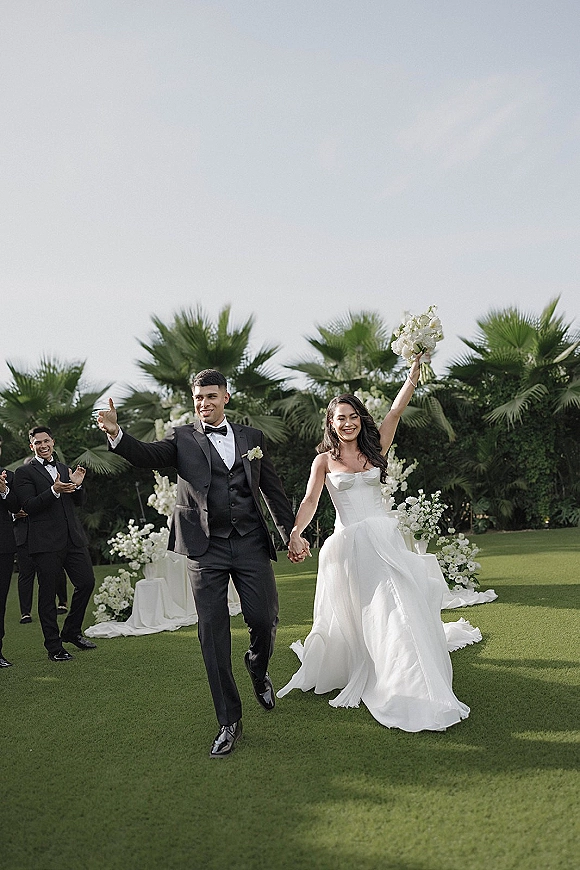 Wedding recessional as bride and groom walking hand in hand, bride raises white bouquet beside floral aisle draping on a palm-lined lawn