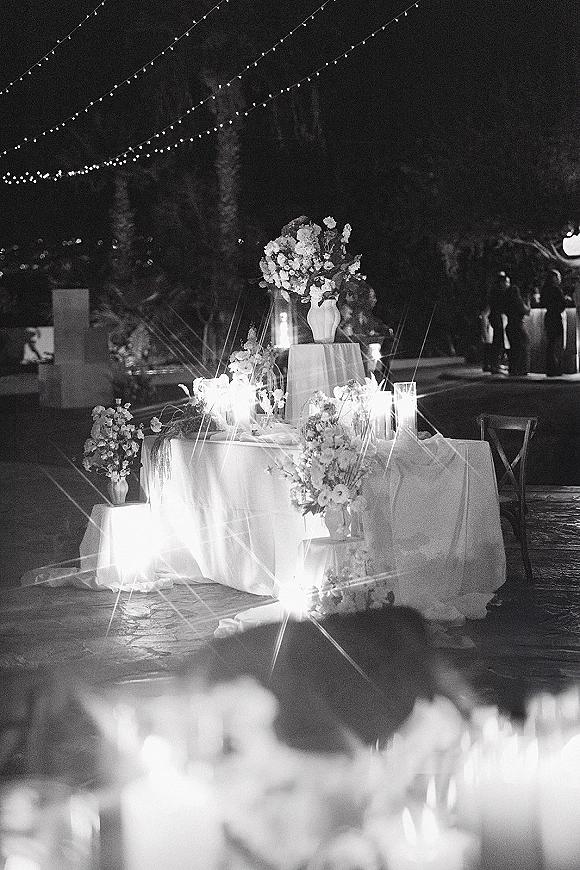 Reception sweetheart table with sweetheart table decor, white linens, floral vases and candles under string lights on an outdoor patio at night