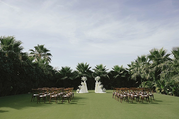 Ceremony setup for an outdoor wedding ceremony with wood crossback chairs in rows, white floral pedestals, and draped fabric on a lawn by palm trees