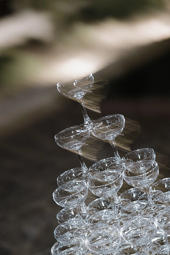 Champagne tower of stacked coupe glasses with shimmering glassware accents glowing under ambient light against a dark blurred background