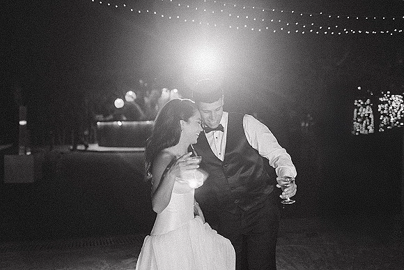 Couple portrait in a black and white wedding photo, bride in strapless dress and groom in vest laughing with cocktails under bistro lights at night