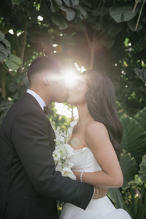 Wedding kiss portrait with sun flare as bride in strapless dress holds white bouquet, kissing groom in tux amid lush tropical greenery