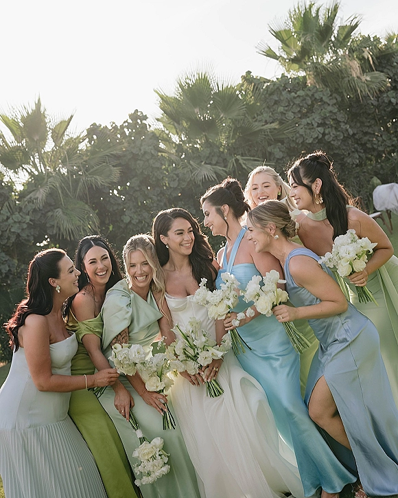 Bridesmaid group photo of bride with bridesmaids laughing, holding white bouquets in pastel dresses under palm trees in bright garden sky