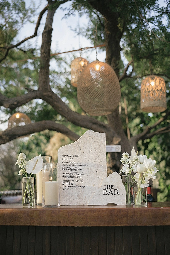 Wedding bar signage on a stone bar sign with glass cylinder candles, white flowers, wine bottles, and wicker lanterns over an outdoor bar in trees