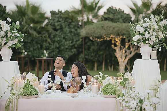 Sweetheart table moment with bride and groom laughing amid white florals and candles, set on a lawn with palm trees and string lights