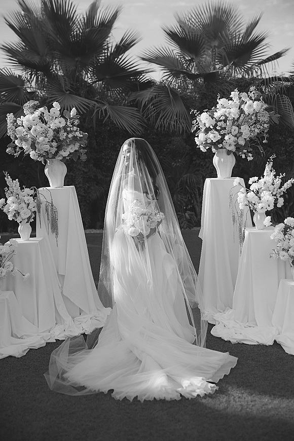 Bridal portrait of a bride in a long veil holding a white bouquet, standing between floral pedestal arrangements in a palm-lined garden setting