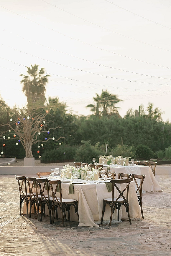 Reception tablescape at an outdoor wedding reception with long white linen tables, cross-back chairs, white floral centerpieces, and string lights on a stone patio