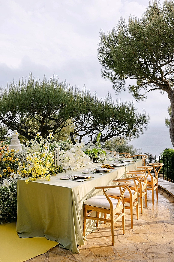 Reception tablescape with long banquet table setup on a green tablecloth, yellow-white florals, and cake on an ocean-view terrace