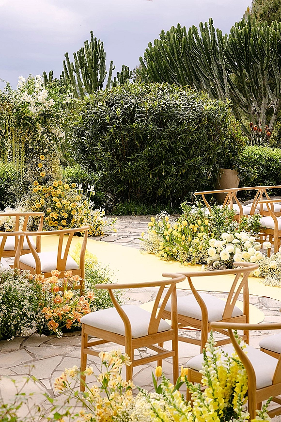 Ceremony aisle decor with a yellow wedding aisle runner, wooden chairs with white cushions, and low white and yellow florals on a stone patio