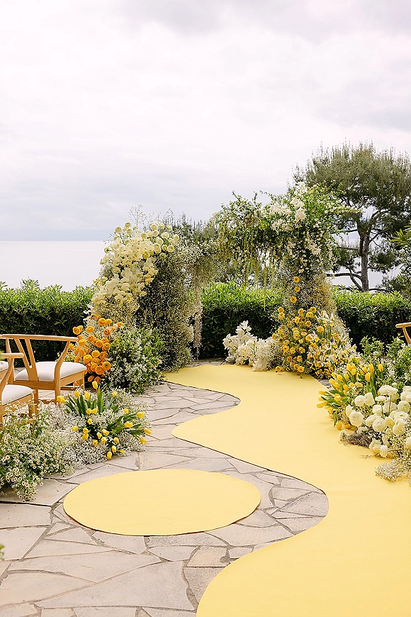 Ceremony aisle decor with a curved yellow wedding aisle runner, meadow-style flowers, and a hanging floral arch on a stone patio by the ocean view