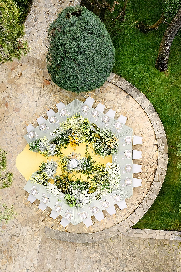 Reception tablescape with an overhead wedding tablescape view of a curved banquet table, yellow linens, white florals, and taper candles on a stone patio