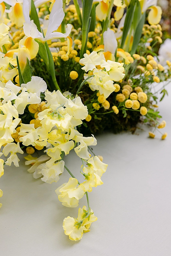 Wedding centerpiece with yellow and white flowers and lush green foliage arranged low on a white tabletop, garden-style and airy