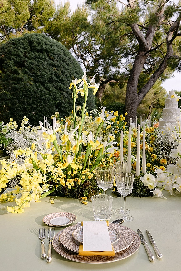 Reception tablescape with yellow and white floral centerpiece, taper candles, crystal stemware, and patterned chargers in an outdoor garden setting