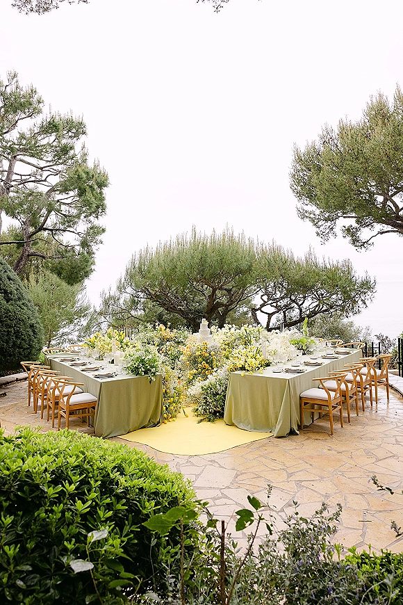 Reception tablescape with outdoor reception tables in a U-shape, green tablecloths, floral centerpieces, candles, and tiered cake on a stone patio